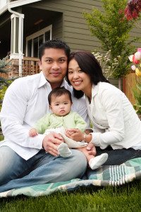 Husband and Wife Hold Their Baby while Sitting on a Blanket in Front of Their House and Smiling at the Camera